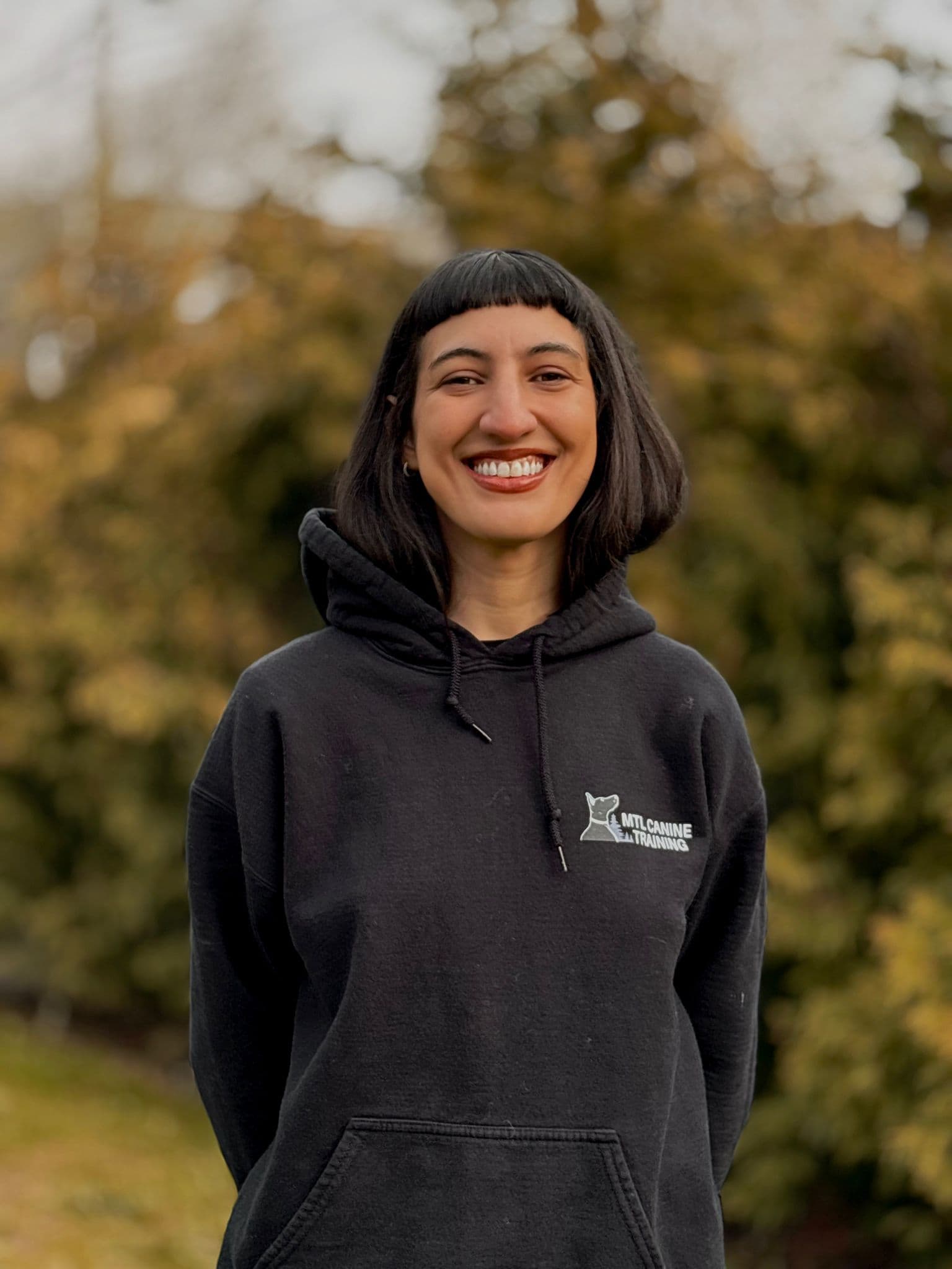 Portrait de Mia M souriante, vêtue d'un sweat à capuche « Montreal Canine Training », en plein air